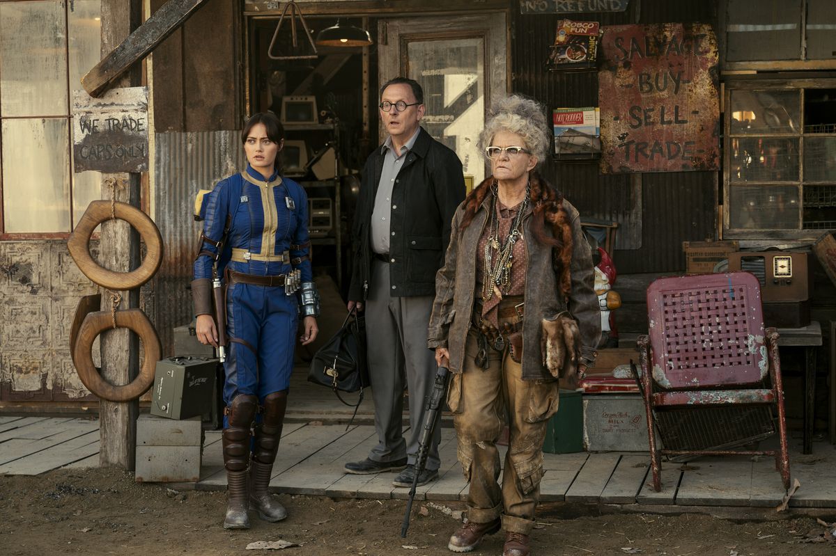 Lucy (Ella Purnell) stands with a scientist (Michael Emerson) and a shopkeeper from the Wasteland (Dale Dickey) all looking at something