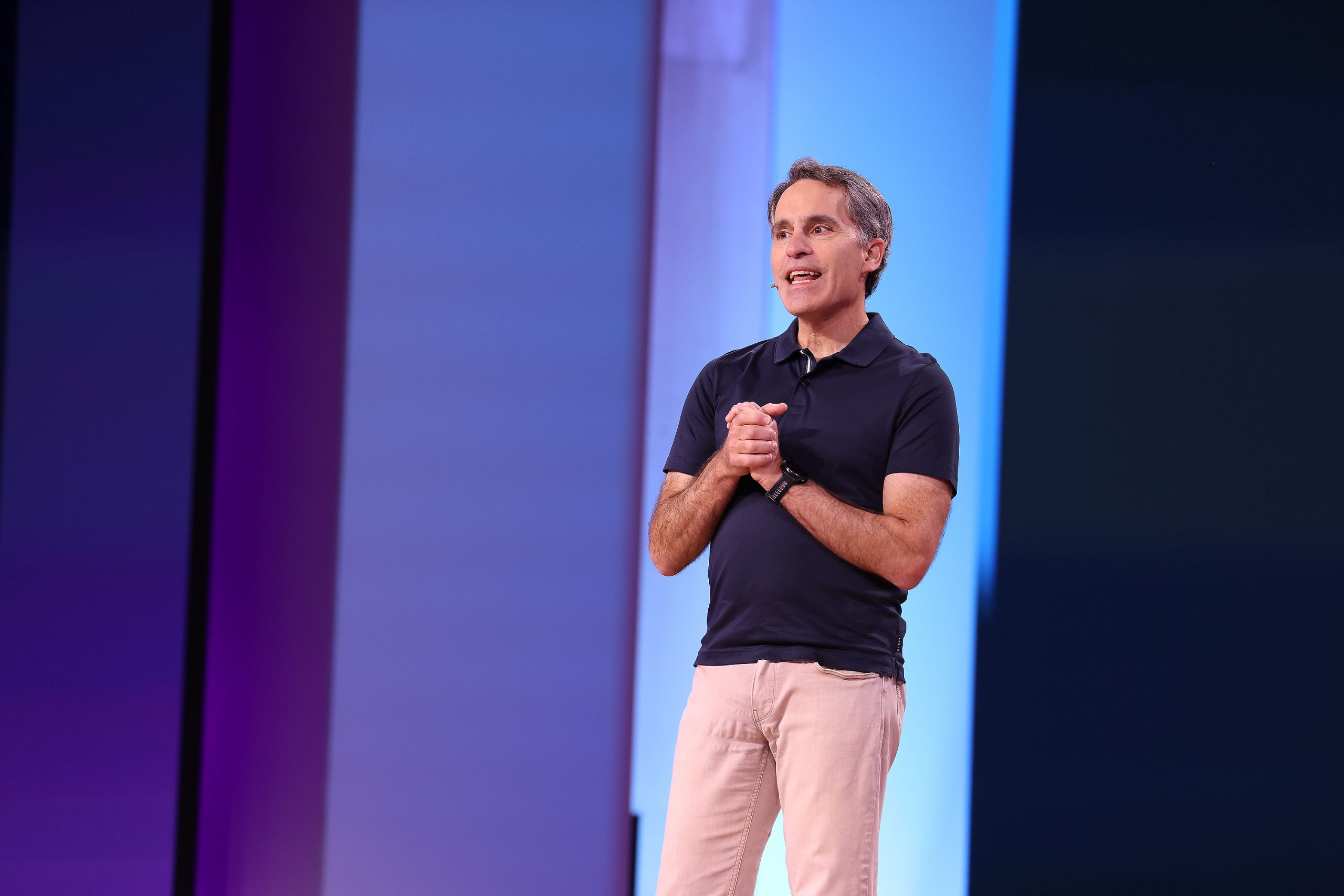 A man standing on stage with his hands clasped and a blue and purple screen behind him