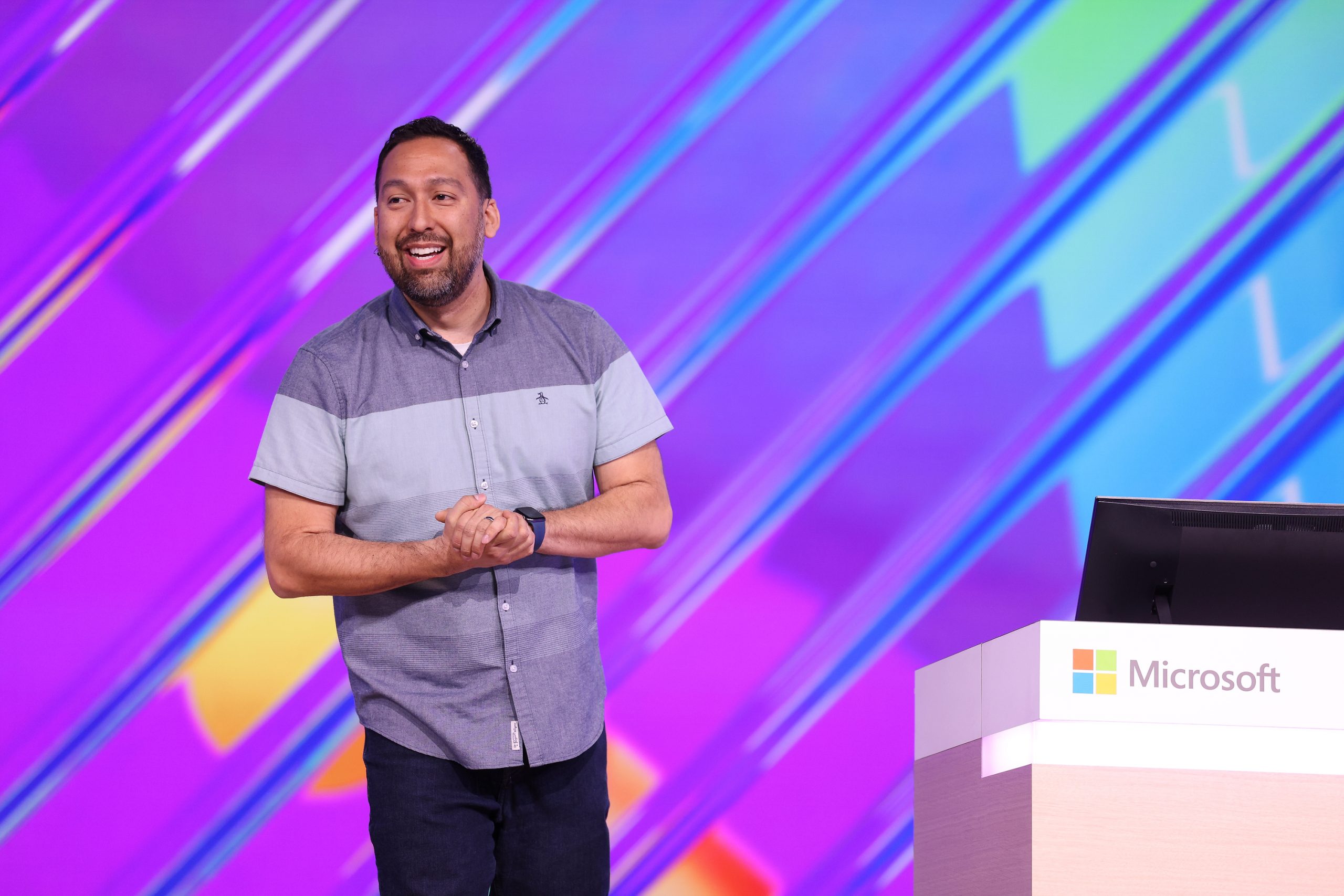 A man standing on stage speaking to an audience with his hands clasped and a table and laptop beside him