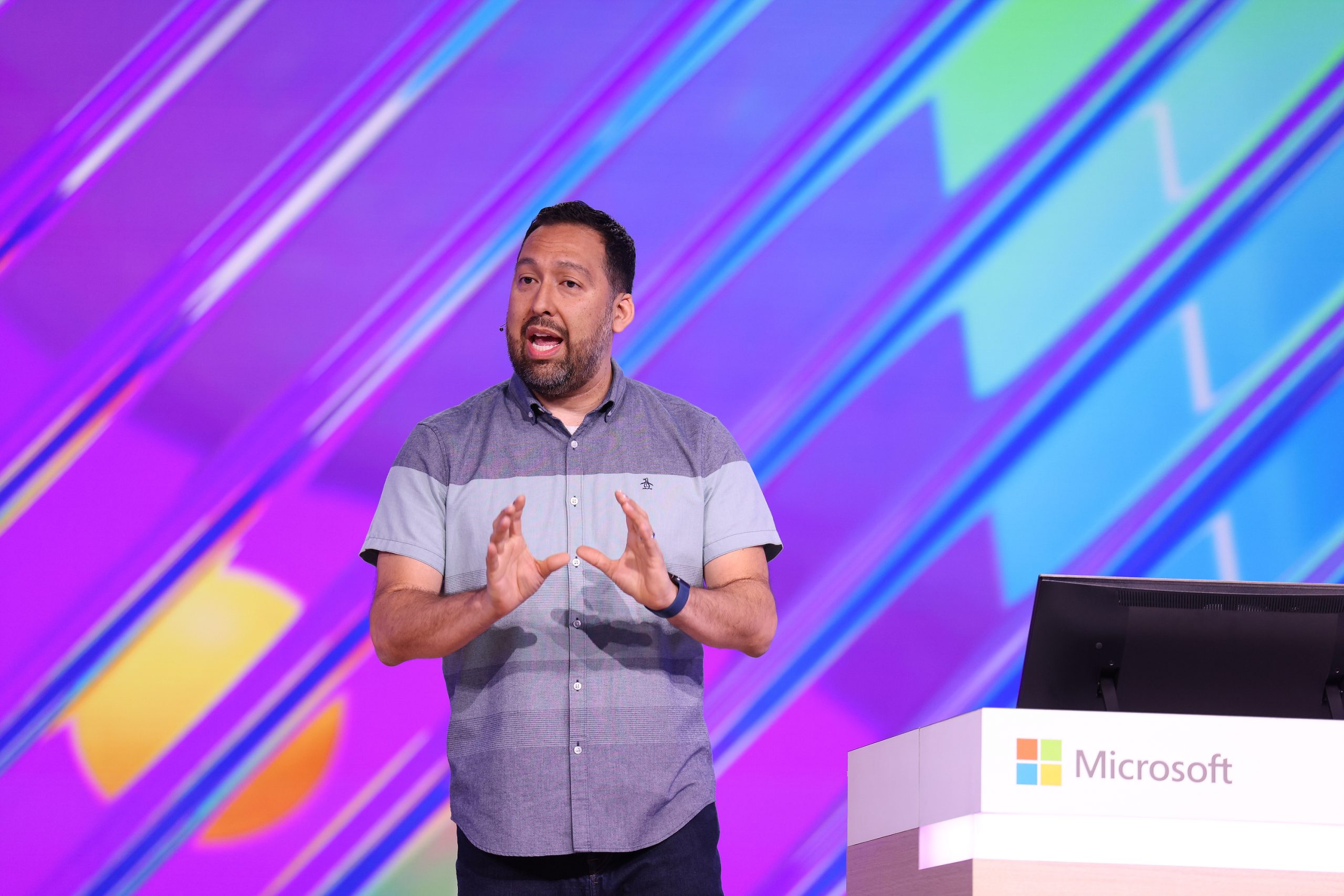 A man standing on stage speaking to an audience and gesturing with his hands with a table and laptop beside him