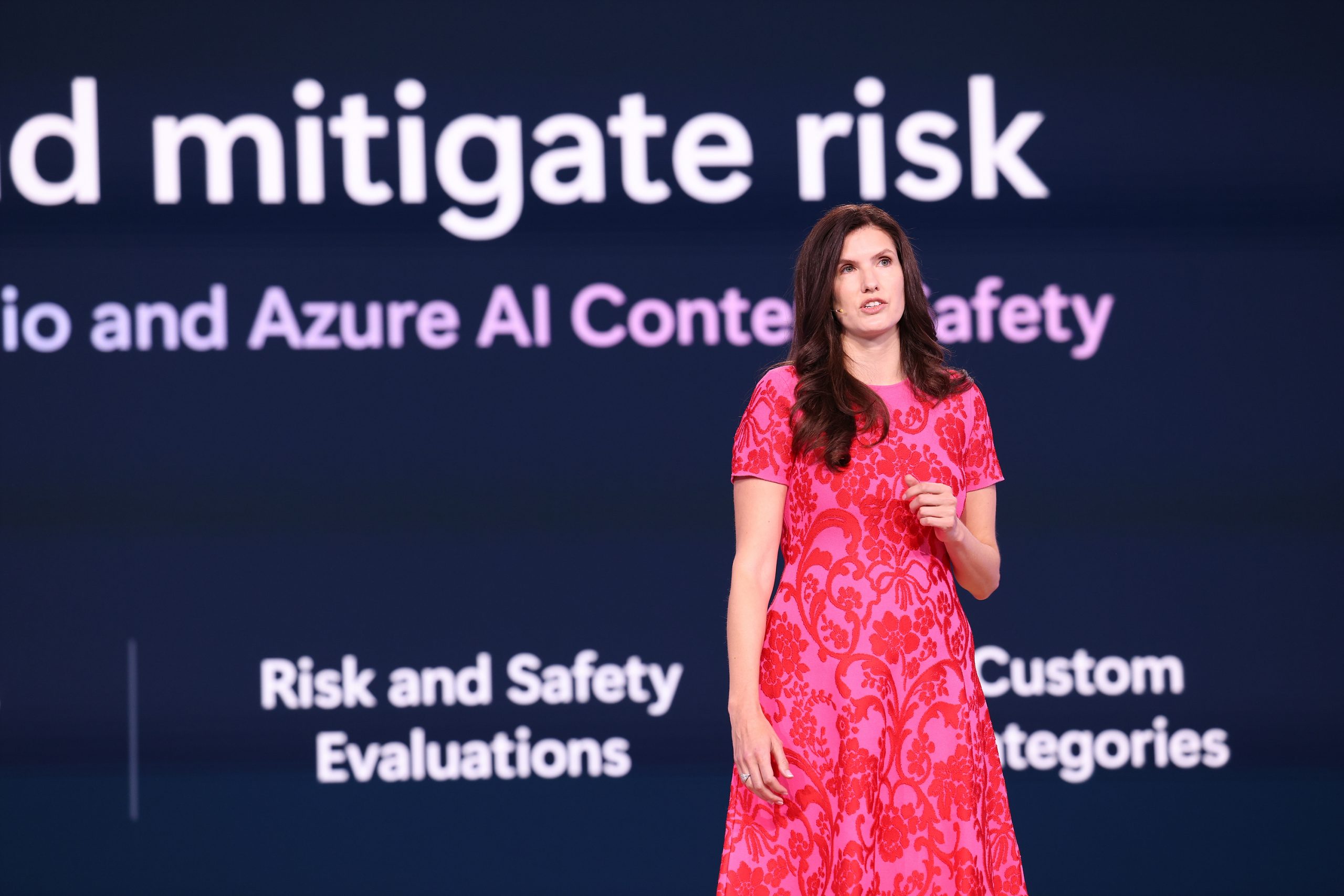 A woman standing on stage speaking to an audience, with text on a screen behind her