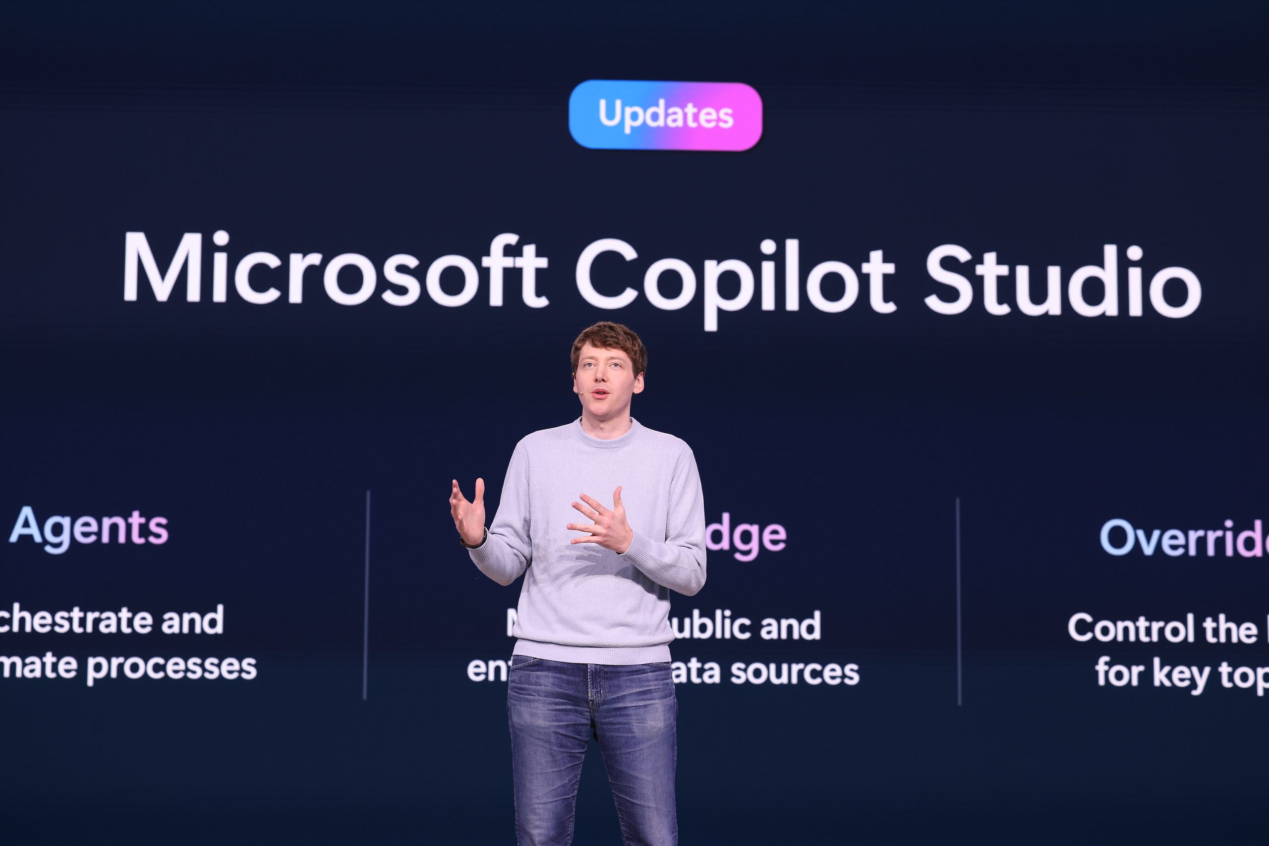 A man standing on stage speaking to an audience and gesturing with his hands, with text on the screen behind him about Microsoft Copilot Studio